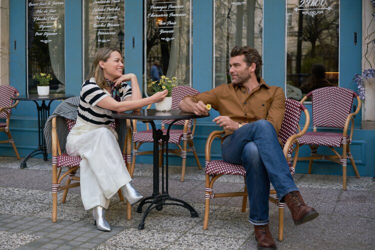 A man and a woman sitting outside a café, sharing a lighthearted moment. The woman is smiling and leaning towards the man, who is holding a flower.