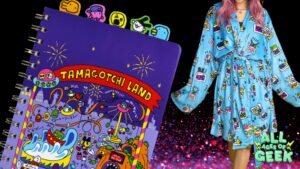 The image showcases a vibrant Tamagotchi-themed spiral notebook and a robe. The notebook, titled "Tamagotchi Land," features colorful amusement park illustrations with Tamagotchi characters and fireworks. The robe, worn by a person with pink hair, is light blue with various Tamagotchi characters and devices printed on it. The "All Ages of Geek" logo is displayed in the bottom right corner.