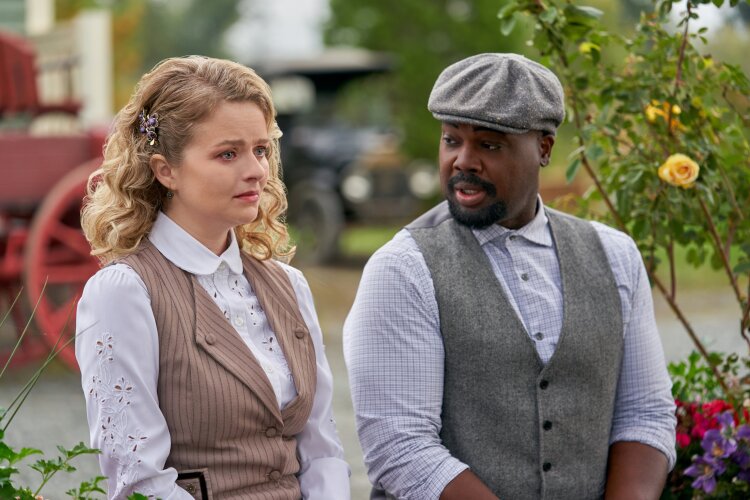 A woman with curly blonde hair and a man wearing a flat cap and vest sitting outdoors, having a serious conversation. From Hallmark's Truth Be Told When Calls the Heart.