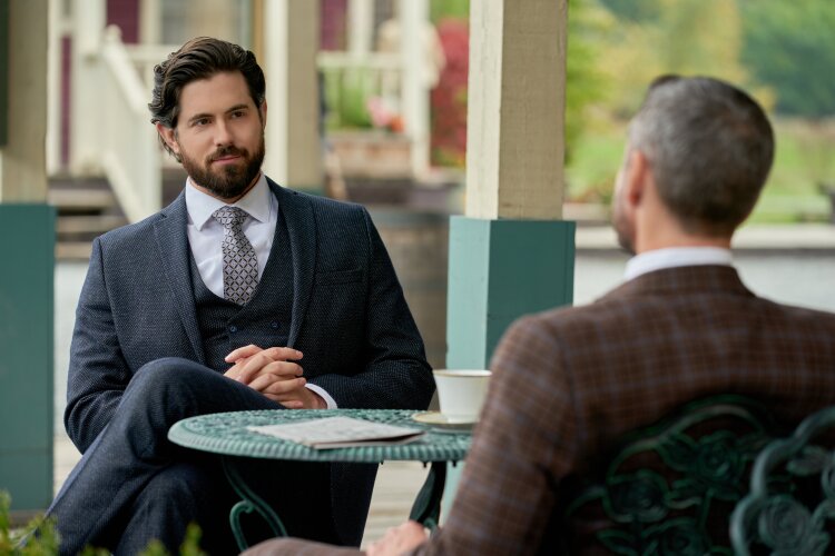 A man with dark hair and a beard, wearing a dark suit, sitting at a table with another man in a checkered suit, talking over coffee. From Hallmark's Truth Be Told When Calls the Heart.