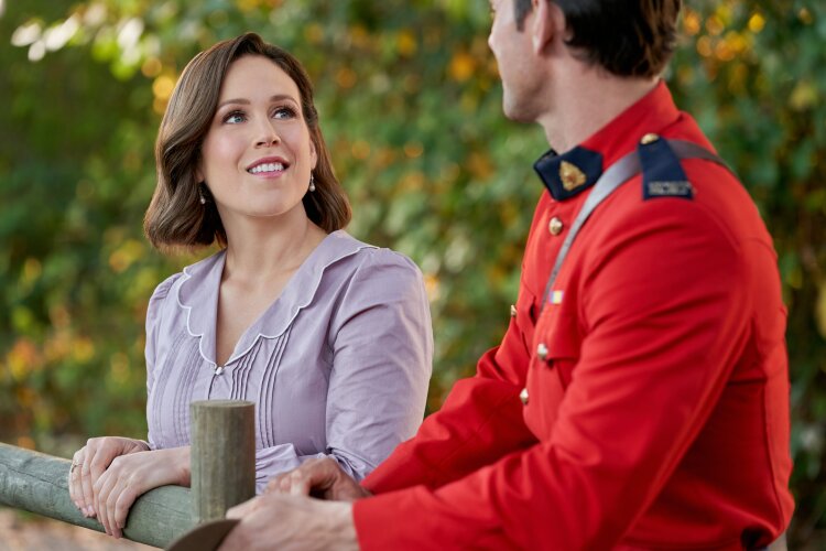A woman with short brown hair in a light purple blouse, smiling while talking to a man in a red uniform, outdoors with greenery in the background.
