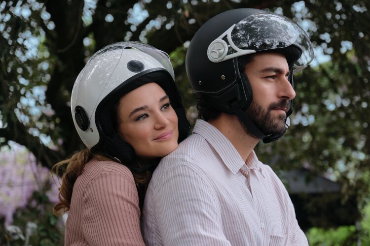 A woman and man wearing helmets, sitting on a parked scooter, with the woman hugging the man from behind.