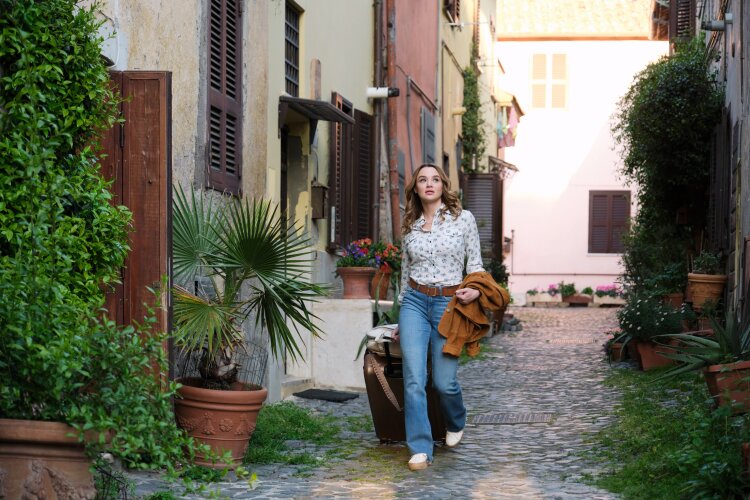 A woman walking down a charming, narrow alleyway in Italy, carrying a suitcase and a jacket.