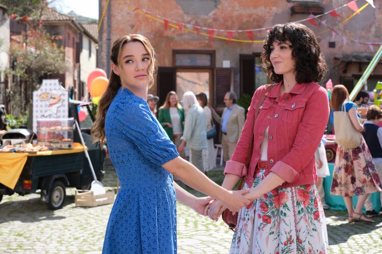 Two women holding hands at a festive outdoor event, surrounded by people and decorations.  "Two Scoops of Italy"  on Hallmark.