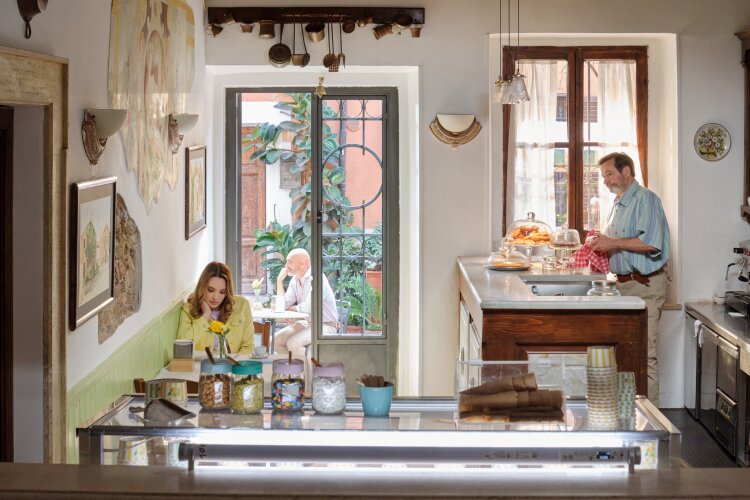 A bright and inviting Italian café with a woman sitting by the window, reading a book, and an older man working behind the counter.  "Two Scoops of Italy"  on Hallmark.