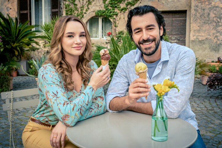 A smiling man and woman sitting outside, enjoying ice cream cones at a small table with a vase of flowers.