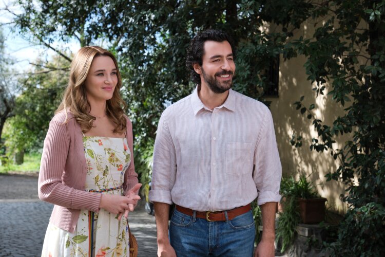 A couple standing side by side, smiling, and looking towards something off-camera, in a green and sunny outdoor setting.