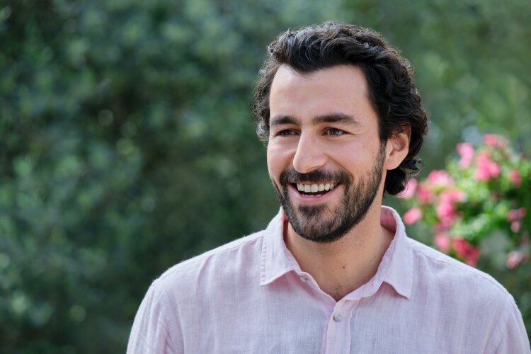 A close-up of a man with curly hair and a beard, smiling warmly with a backdrop of greenery and flowers.  "Two Scoops of Italy"  on Hallmark.