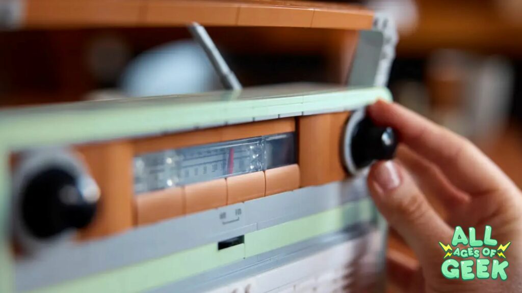 ### Alt Text for Image A close-up of a hand turning the dial on a LEGO® replica of a 1970s transistor radio. The build features detailed design elements, including a textured grille and authentic-looking buttons and dials. The logo for "All Ages of Geek" is visible in the bottom right corner of the image.