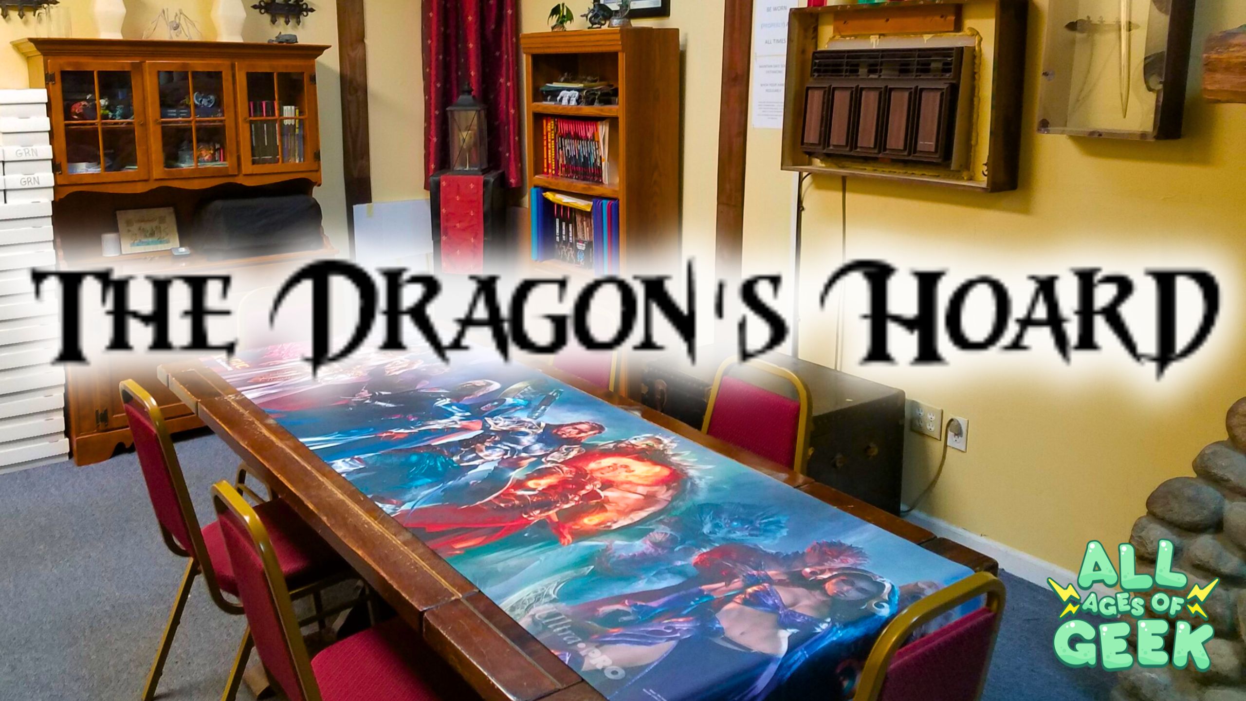 The image shows the interior of a game store with a large table in the center covered with a colorful fantasy-themed playmat. The background features shelves filled with books and gaming supplies, and there's a wooden cabinet displaying various items. The text "The Dragon's Hoard" is prominently displayed in the foreground, and the "All Ages of Geek" logo is located at the bottom right corner. The overall atmosphere is cozy and inviting, perfect for gamers.