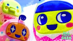 Close-up photo of three colorful Tamagotchi plush toys against a bright sky background. The plushies resemble classic Tamagotchi devices with cheerful faces and vibrant colors. One plush is lime green, another is pink with a character featuring starry eyes, and the third is yellow with a blue face and large, expressive eyes. The "All Ages of Geek" logo is displayed at the bottom right corner.