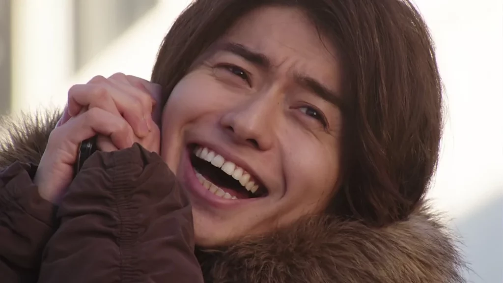 Kazumi Sawatari from Kamen Rider Build, smiling joyfully with his hands clasped together, wearing a fur-lined jacket.