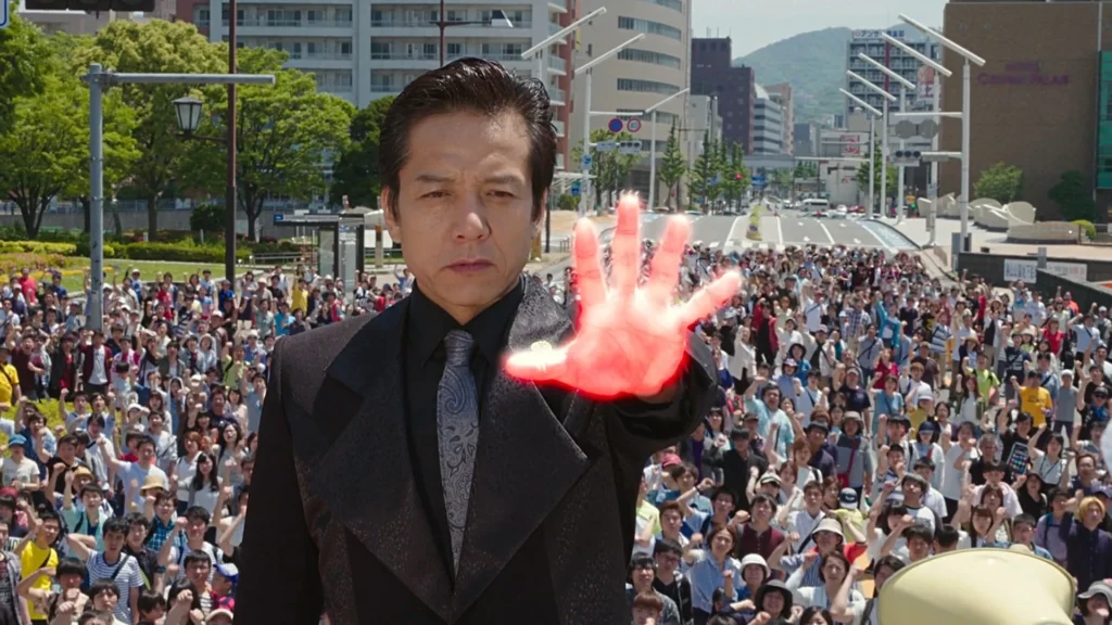 Kengo Ino from Kamen Rider Build, standing in front of a large crowd, holding out his glowing red hand, wearing a black suit and tie, with a serious expression.
