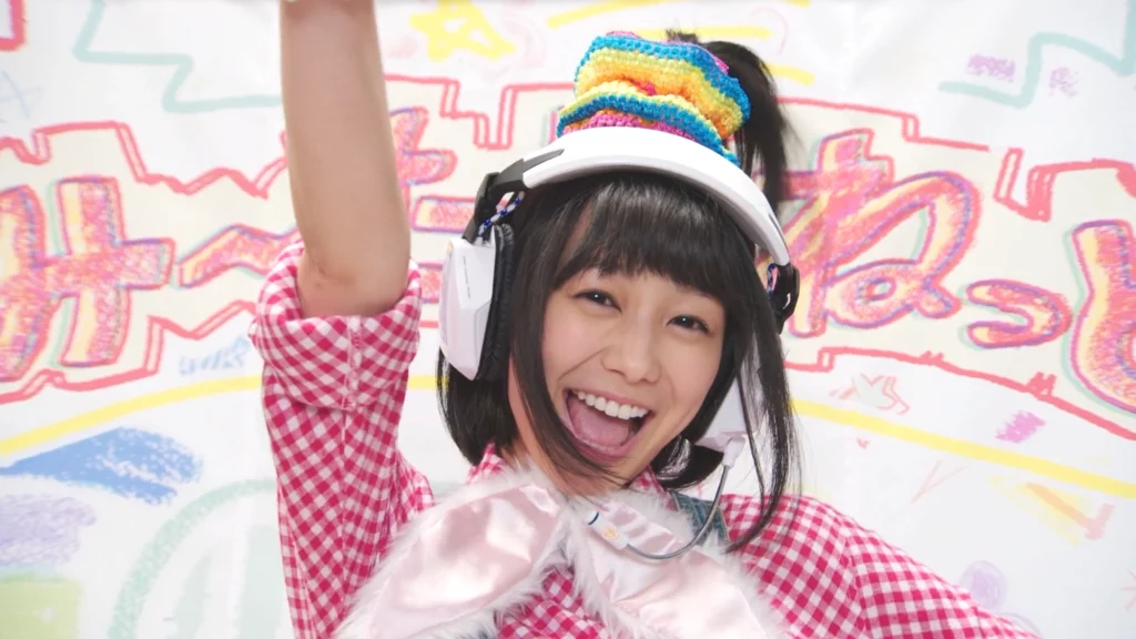 Misora Isurugi from Kamen Rider Build, smiling brightly with her arm raised, wearing a colorful headband and headphones, in front of a colorful background.
