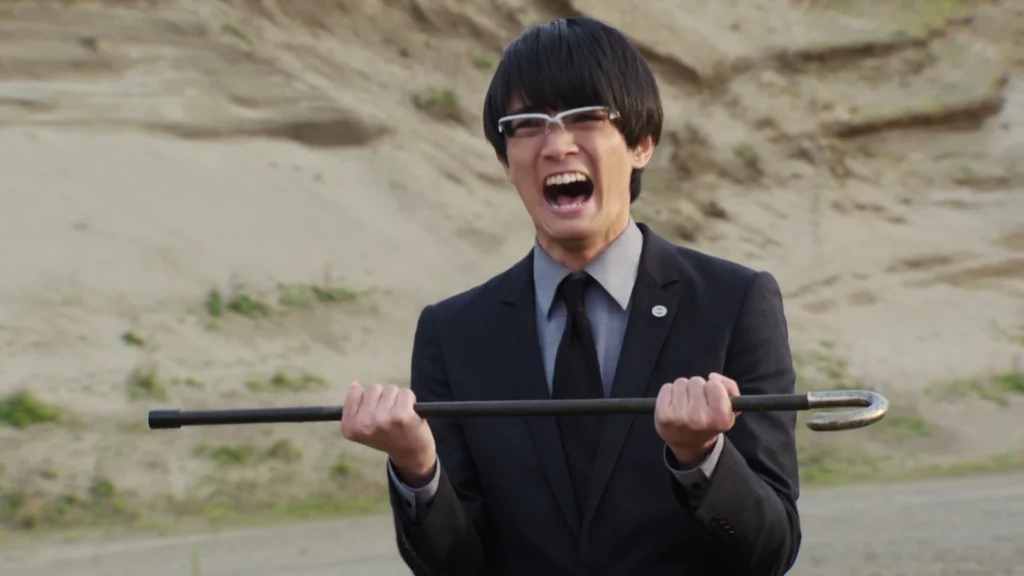 Nariaki Utsumi from Kamen Rider Build, wearing a suit and glasses, holding a metal rod, and shouting with intense emotion in a desert-like environment.