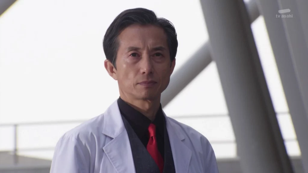 Shinobu Katsuragi from Kamen Rider Build, wearing a white lab coat over a black shirt and red tie, with a serious expression and a plain background.