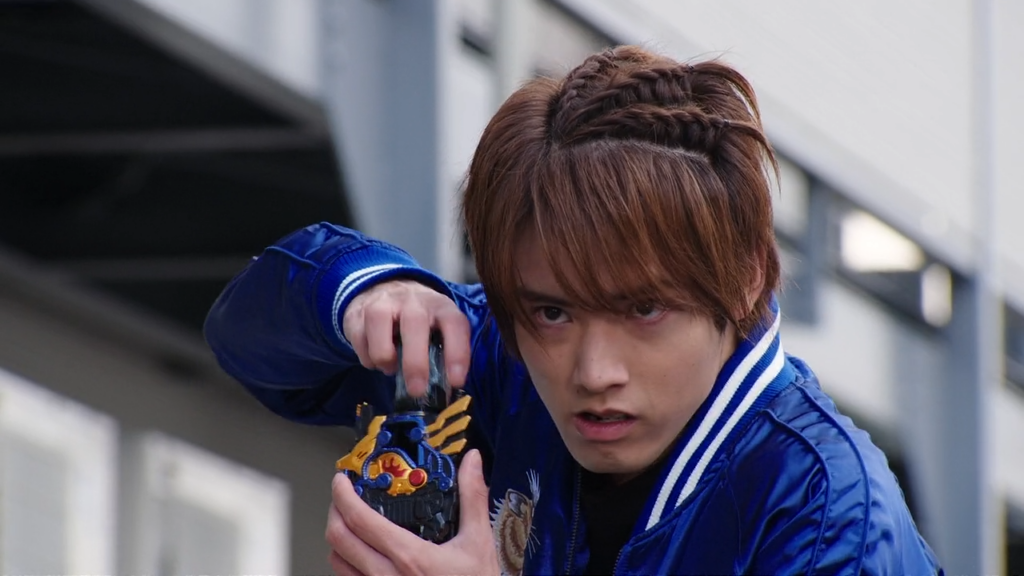Ryuga Banjou from Kamen Rider Build in a determined pose, holding a transformation device, wearing a blue jacket and looking focused.