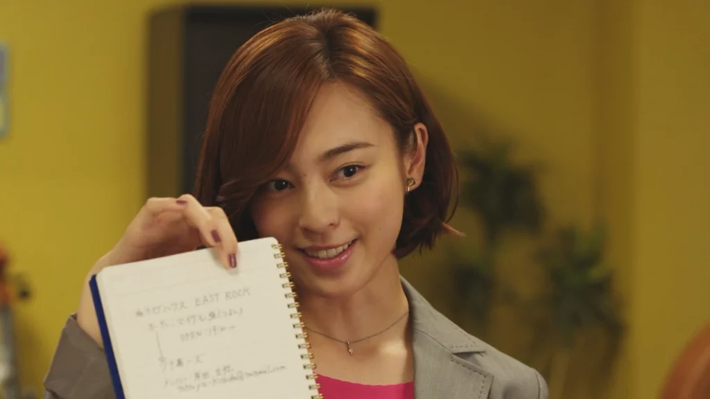 Sawa Takigawa from Kamen Rider Build, smiling and holding up a notebook with notes, dressed in a grey blazer, in a warmly lit room.