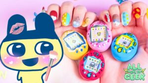 A colorful promotional image for the "Tamagotchi Lip & Cheek" cosmetics. The background features a pair of hands with bright, multi-colored, and intricately designed nails holding four miniature Tamagotchi devices in pink, yellow, and blue. Each Tamagotchi has a retro digital pet displayed on its screen. On the left side, there's an enlarged cute Tamagotchi character with a pink kiss mark on its head and holding a makeup brush. The All Ages of Geek logo is displayed in the bottom right corner. The overall design is vibrant and playful, reflecting the nostalgic and fun essence of Tamagotchi.