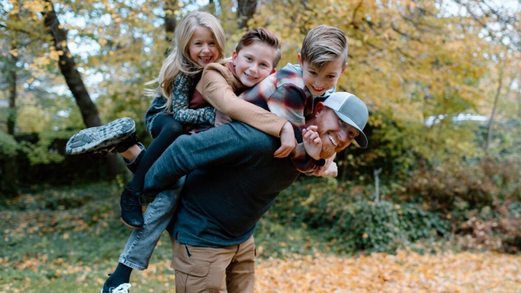 A joyful family moment captured outdoors in the autumn. A father, wearing a gray cap and sweater, carries three smiling children on his back. The two boys, one in a plaid shirt and the other in a brown jacket, and a girl with long blonde hair, are dressed warmly for the fall weather. The vibrant, golden leaves on the ground and trees create a beautiful seasonal backdrop, reflecting the joy and warmth of spending quality family time together.
