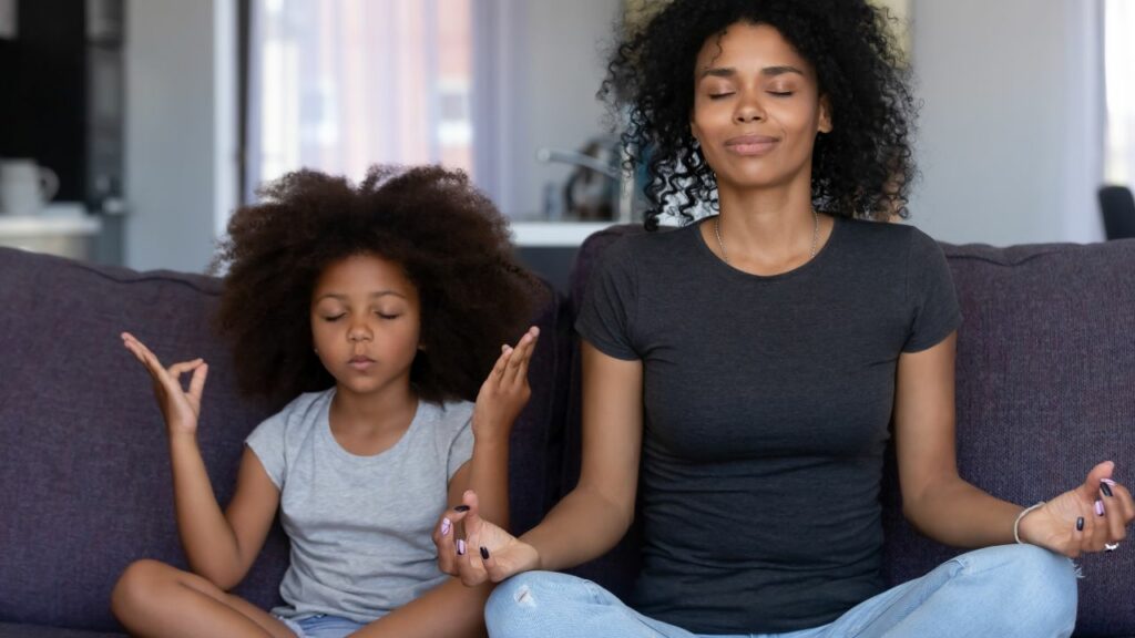 A serene moment of mindfulness captured at home. A mother and her young daughter sit cross-legged on a cozy sofa, eyes closed, deeply engaged in meditation. Both wear comfortable, casual clothing, the mother in a dark t-shirt and the daughter in a light gray one, exuding a sense of calm and focus. Their peaceful expressions and the soft lighting create a warm, inviting atmosphere, highlighting the importance of taking time for mental well-being in the midst of daily routines.
