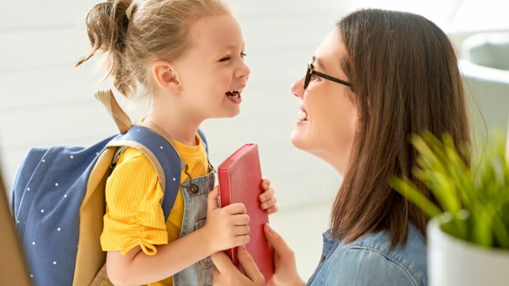 A joyful moment of connection in homeschooling. A mother, dressed casually in a denim shirt and wearing glasses, shares a hearty laugh with her young daughter. The child, with her hair tied up and dressed in a bright yellow shirt and overalls, carries a blue backpack and holds a red book. Their faces are lit with happiness, capturing the essence of open communication and the strong bond in their homeschooling journey. The warm, inviting setting with soft lighting enhances the feeling of a supportive and nurturing learning environment.