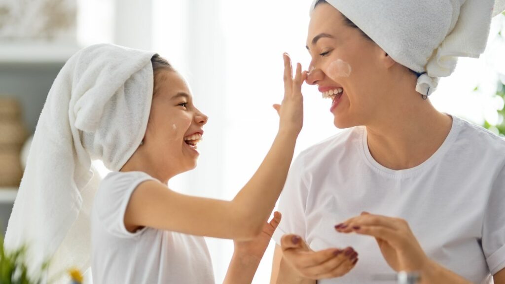 A delightful moment of self-care and bonding in homeschooling. A mother and her young daughter share a joyful laugh while applying skincare products. Both have white towels wrapped around their heads, enhancing the spa-like atmosphere at home. The mother, dressed in a simple white t-shirt, smiles warmly as her daughter gently applies lotion to her nose. The bright and airy setting, with soft natural light, underscores the importance of practicing self-care and spending quality time together, fostering a nurturing and supportive homeschool environment.