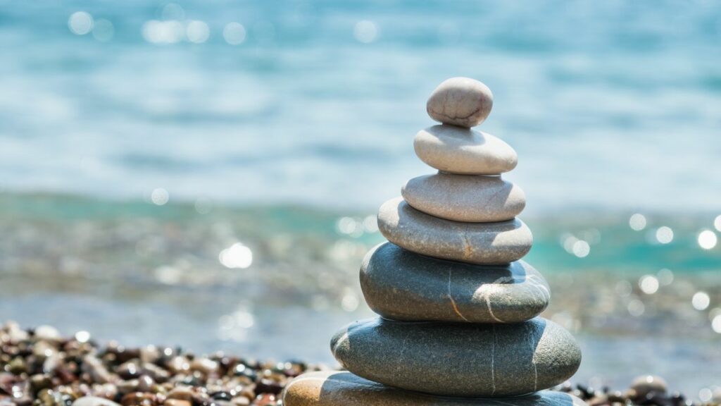 A serene image showcasing the practice of mindfulness and relaxation techniques, perfect for homeschooling environments. Smooth stones are carefully balanced on a beach, creating a peaceful tower against the backdrop of a calm, blue sea. The gentle waves and sparkling water evoke a sense of tranquility and balance, highlighting the importance of incorporating moments of relaxation and mindfulness into the homeschooling routine. This visual reminder encourages taking time to breathe, center oneself, and find harmony amidst the daily educational activities.