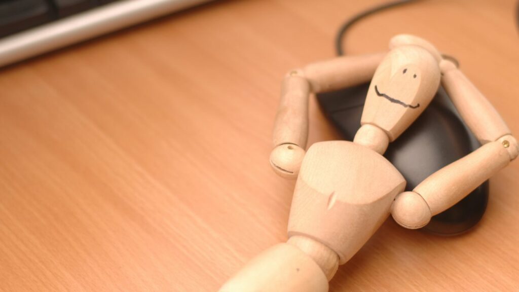 A playful image illustrating the importance of scheduling breaks in a homeschooling routine. A wooden mannequin, smiling and relaxed, lies back against a computer mouse, symbolizing the need to take a pause from screen time and structured activities. This visual reminder encourages incorporating regular breaks into the day to recharge and refresh, ensuring a balanced and effective learning experience for homeschooling families.