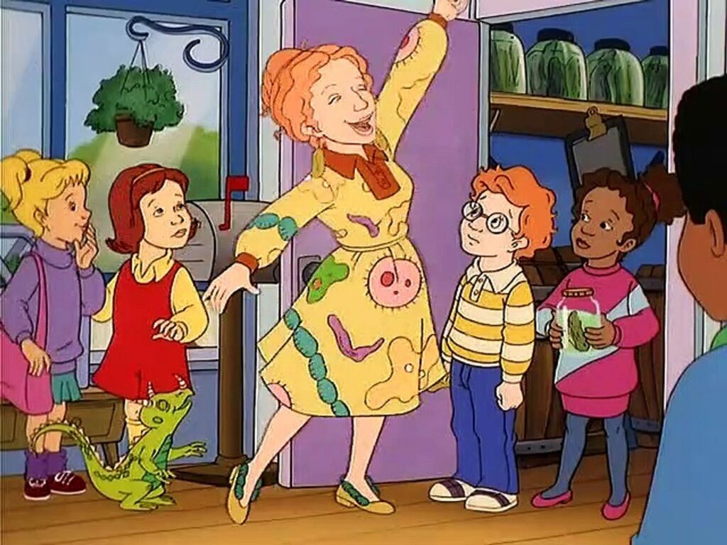 An animated scene featuring Ms. Frizzle, the red-haired teacher from 'The Magic School Bus,' joyfully posing in her colorful dress adorned with science-themed patterns. Surrounding her are four students with varied expressions, including a girl in a purple hoodie, a girl in a yellow shirt and red dress, a boy with glasses in a yellow striped shirt, and a girl in a pink and blue sweater holding a jar with a specimen. A green lizard is on the floor near Ms. Frizzle. They are inside a classroom with shelves and jars visible in the background.