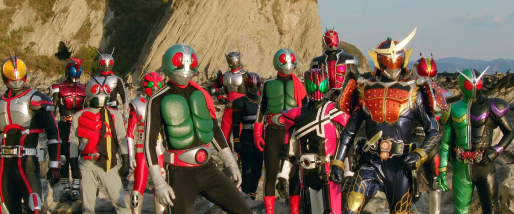 A group of Kamen Riders from various eras, featuring both Showa and Heisei Riders, stand together on a rocky terrain. The lineup includes classic designs like Kamen Rider 1 with his green armor and red scarf, alongside more modern Riders like Kamen Rider Ex-Aid, Kamen Rider Decade, and Kamen Rider Gaim, with their intricate and colorful suits. The newer Riders are distinguished by their sleeker, more complex armor and vibrant colors, while the older Riders retain their retro, minimalist designs. The scene captures a fusion of past and present Kamen Rider heroes united.