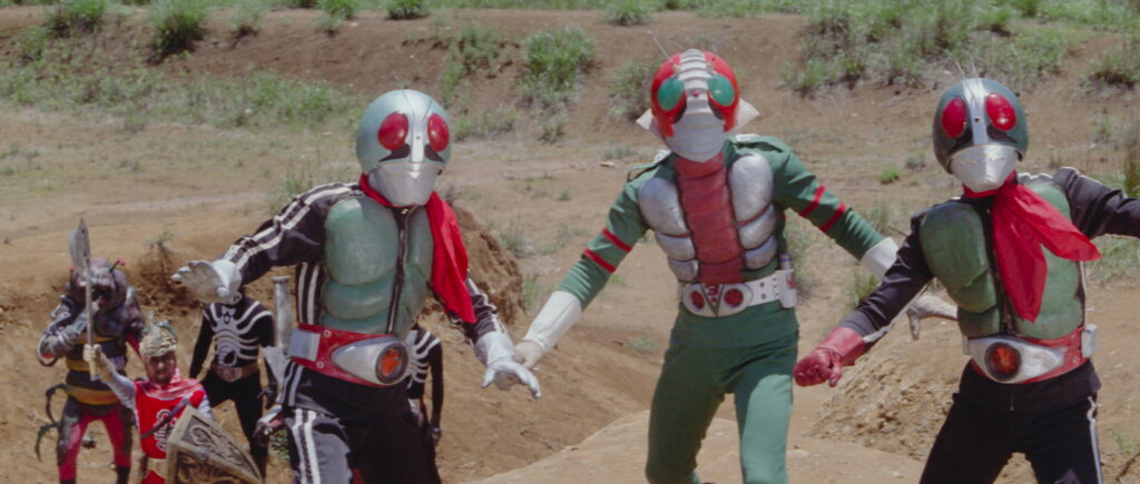 Three Kamen Riders in classic Showa-era suits, wearing green armor, red scarves, and bug-eyed helmets, stand in a battle-ready pose. They are joined by enemies in the background, set against a barren outdoor landscape with grass patches. The middle Rider, possibly Kamen Rider V3, wears distinct red and green armor with a white belt.