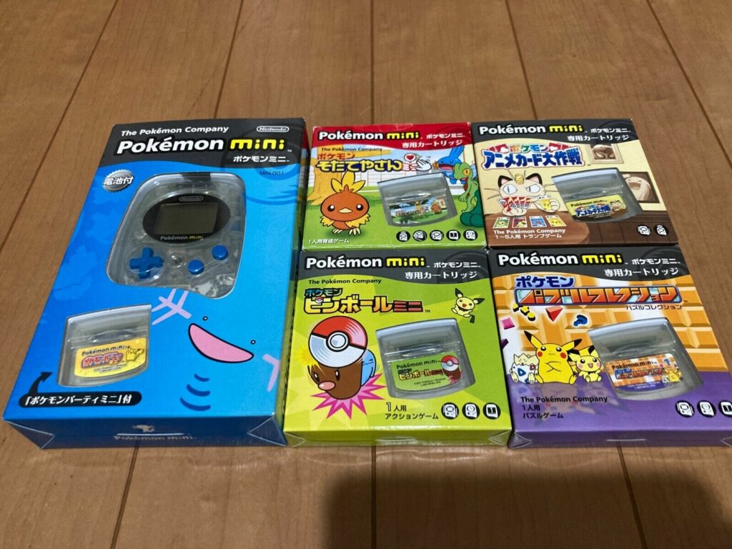 A boxed Pokémon Mini handheld console in blue, featuring Wooper on the packaging, is displayed alongside four Pokémon Mini game cartridges. Each cartridge comes in its own box with unique cover art: one features Torchic, another shows a Poké Ball, while others highlight Pikachu and various Pokémon-themed mini-games. The games included are Pokémon Party Mini, Pokémon Pinball Mini, Pokémon Puzzle Collection, and more, showcasing a variety of puzzle and action-based Pokémon games. All items are neatly laid out on a wooden surface, giving a nostalgic and collectible vibe.