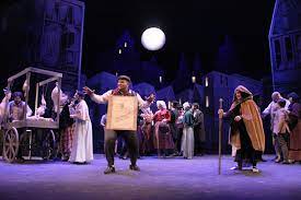 A theatrical production set under a full moon features a bustling town square scene, with performers dressed in period costumes engaging in lively activity. A man in the foreground commands attention, holding a prop or sign, while others around him add depth and drama to the scene, creating a vibrant and immersive stage atmosphere.