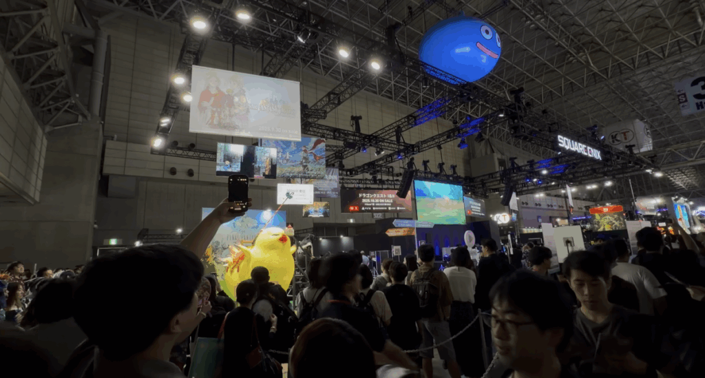 Crowds gather at the Dragon Quest booth during Tokyo Game Show 2025, featuring a giant inflatable blue Slime hanging above, large video screens, bright lights, and colorful displays across the bustling convention floor.