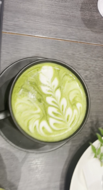 A beautifully crafted matcha latte with intricate leaf-style latte art served in a black cup on a dark wooden table. From Iyemon Salon Umeda.