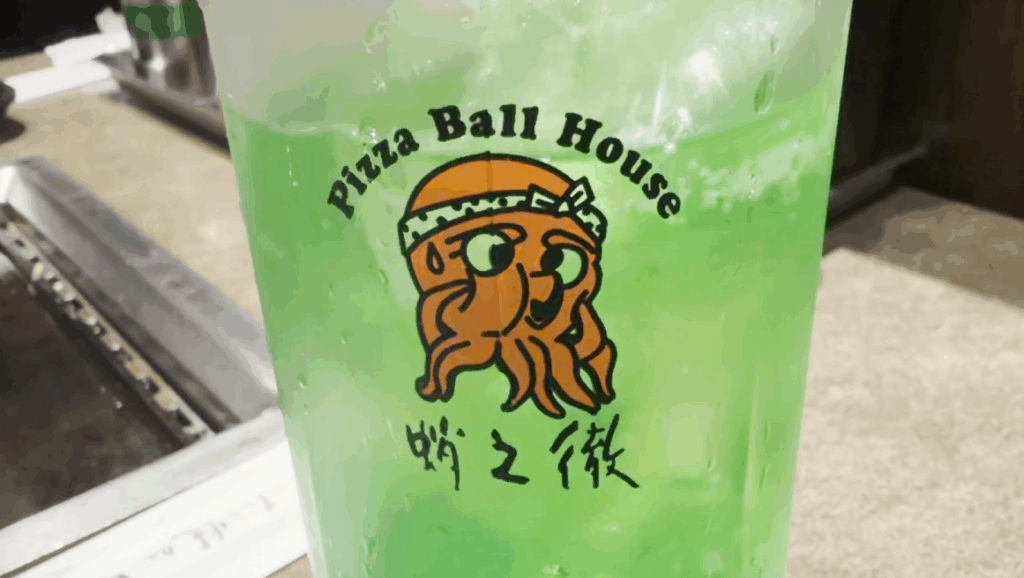 A glass of bright green melon soda with the Pizza Ball House logo, served at Takonotetsu Tsunoda in Osaka.