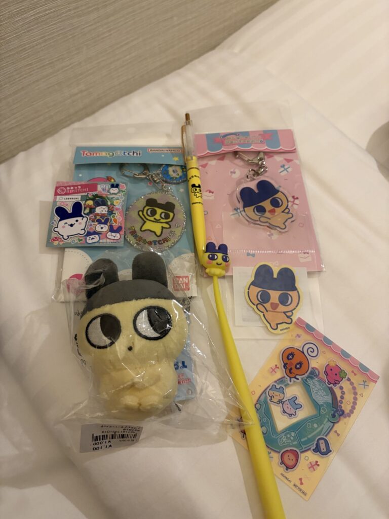 A collection of Tamagotchi merchandise including plush toys, keychains, a pen, and stickers featuring classic characters displayed on a hotel bed.